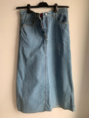 ICHI Lång jeanskjol i blå denim - Snygg lång jeanskjol i klassisk blå denim med hög midja, framfickor och dragkedja framtill. Kjolen har bälteshällor och raka linjer för en clean look. Perfekt att styla med sneakers eller boots.