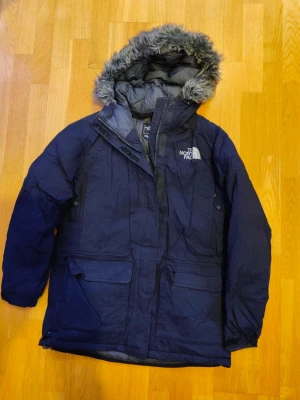 Mörkblå dunjacka från The North Face - Mörkblå dunjacka från The North Face med huva och grå fuskpäls. Jackan har två stora fickor framtill, dragkedja och tryckknappar. Perfekt för kalla vinterdagar. Herrjacka. Inga byten.