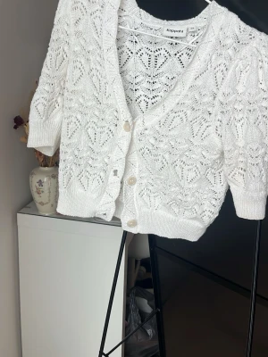 Vit virkad kofta från Kappahl - Söt vit kofta från Kappahl med virkade detaljer och korta puffärmar. Koftan har v-ringning, knappar framtill och ett fint spetsmönster som ger en romantisk känsla. Perfekt att slänga över en topp för en trendig look.
