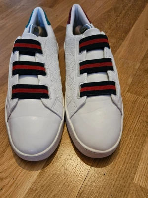 Gucc Ace vita sneakers med färgglada detaljer - Säljer ett par vita Gucci Ace sneakers i läder med klassiska gröna och röda ränder på elastiska band istället för snören. Baksidan har metallicdetaljer i rött på ena skon och grönt på den andra. Diskret GG-mönster i lädret ger extra lyxig känsla.