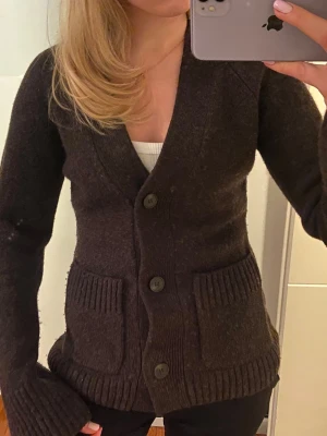 Wera brun stickad kofta med fickor - Mysig brun stickad kofta från Wera med stora knappar och två rejäla fickor framtill. Koftan har långa ärmar med breda ribbade muddar och en djup v-ringning. Perfekt att slänga på sig när det är lite kyligt.