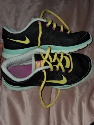 Nike svarta sneakers med gula detaljer - Snygga svarta Nike sneakers med gula skosnören och swoosh-logga. Skorna har mintgröna sulor och lila innersulor med rosa Nike-tryck. Lågt skaft och meshmaterial för extra komfort. Perfekta för dig som vill sticka ut med färgglada detaljer. Storlek 35,5. Även tvättade. De har lite lödder innuti