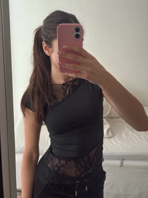 Svart topp med spetsdetaljer - Snygg svart topp med korta ärmar och asymmetriska spetsdetaljer upptill och nedtill. Toppens material är mjukt och stretchigt, och spetsen har ett blommigt mönster som ger en elegant touch. 