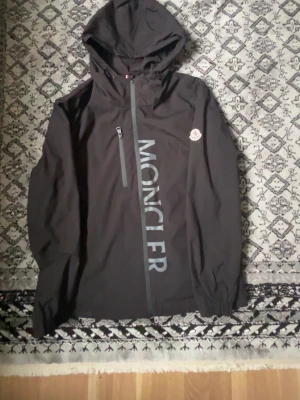 Svart Moncler vindjacka med huva - Snygg svart vindjacka från Moncler med huva och dragkedja framtill. Jackan har ett stort Moncler-tryck på framsidan och logotyp på ärmen. Materialet är lätt och perfekt för blåsiga dagar. Elastiska muddar och diskret tricolor-detalj på ryggen.