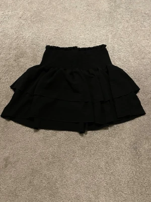 Svart volangkjol med smock - Säljer en svart kortkjol med bred smock i midjan och dubbla volanger. Kjolen har en luftig och flowy känsla, perfekt för att styla med en crop top eller stickad tröja. Materialet känns mjukt och stretchigt, vilket gör den bekväm att bära hela dagen. Köpt på shein och är aldrig använda.