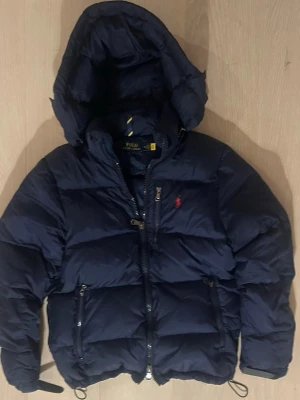 Mörkblå pufferjacka från Polo Ralph Lauren - Snygg mörkblå pufferjacka från Polo Ralph Lauren med röd broderad logga på bröstet. Jackan har huva, dragkedja framtill och två fickor med dragkedja. Perfekt för kalla vinterdagar och riktigt skön att bära. Klassisk design med justerbar nederkant. Det är storlek M men upplevs mer som en S. Saknar en tryckknapp på luvan i övrigt i nyskick. 