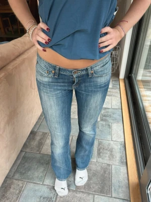 Blå bootcut jeans med låg midja - Säljer ett par blå bootcut jeans med låg midja och slitna detaljer. Jeansen har klassisk femficksmodell och dragkedja med knapp. Perfekta för en avslappnad och trendig look. Materialet är denim i bomull.