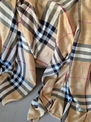 Beige rutig halsduk från Burberry - Klassisk beige halsduk från Burberry i 100% kashmir. Halsduken har det ikoniska rutmönstret i svart, vitt och rött. Tillverkad i Skottland och supermjuk, perfekt för att ge din outfit en lyxig touch.