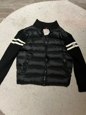 Svart pufferjacka/cardigan från Moncler - Snygg svart pufferjacka från Moncler med stickade ärmar och vita ränder på armarna. Jackan har dragkedja framtill, två fickor med dragkedja och ribbad krage. Moncler-logga på ena ärmen. Perfekt för kalla vinterdagar. Skit fräsch nu inför hösten och varm.