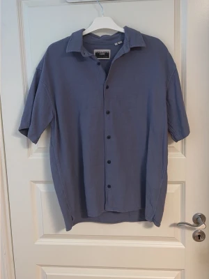 Blå kortärmad skjorta från Jack & Jones - Säljer en stilren blå skjorta från Jack & Jones med korta ärmar och klassisk krage. Skjortan har knappar framtill och är tillverkad i ett mjukt bomullsmaterial. Perfekt för dig som gillar en enkel och avslappnad look.