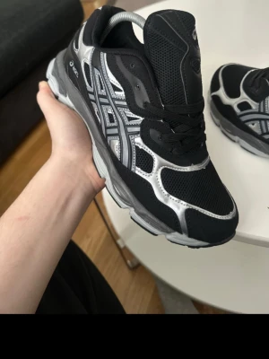 Svarta och silver Asics sneakers - Snygga Asics sneakers i svart och silver med meshpaneler och detaljer i syntetmaterial. Skorna har en sportig design med klassiska Asics-stripes på sidan och tjock sula för extra komfort. Perfekta för dig som gillar streetwear och vill ha en modern look.