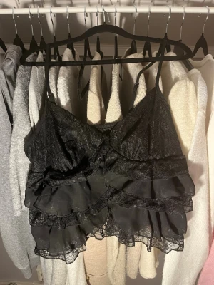 Svart spets topp - Säljer en svart croppad linnetopp med tunna axelband, flera lager volanger och spetsdetaljer. Toppen har en knapp fram och har ett tunnt material, perfekt till sommaren.