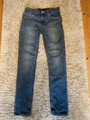 Blå skinny jeans från Levi's - Säljer ett par klassiska blå skinny jeans från Levi's med fem fickor och snygga slitningar framtill. Jeansen har normal midja och en smal passform hela vägen ner. Perfekta till en avslappnad streetstyle-look. Säljer de billigt eftersom att de är för små för mig och för att lappen där bak har lossnat.