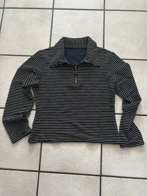 Svartvit randig långärmad topp - Snygg svart långärmad topp med vita tunna ränder och krage. Toppen har en halv dragkedja framtill med en liten ring som detalj. Perfekt till jeans eller kjol för en cool vardagslook.