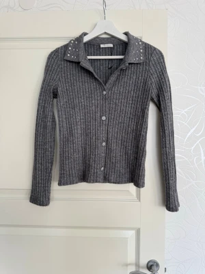 Grå ribbad kofta med pärlor - Snygg grå ribbstickad kofta med knappar framtill och lång ärm. Kragen är dekorerad med pärlor. Står att den är i storlek M men passar mer till S