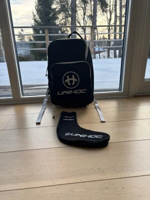 Svart Unihoc ryggsäck med vit logga - Snygg svart ryggsäck från Unihoc med stor vit logga och text på framsidan. Ryggsäcken har vita axelband och flera fack, samt detaljer i slitstarkt tyg. Perfekt för innebandyutrustning eller gymmet. Medföljer ett matchande svart fodral med Unihoc-tryck.