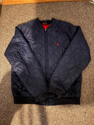 Marinblå bomberjacka Polo Ralph Lauren  - Snygg marinblå bomberjacka från Polo Ralph Lauren med rutit mönster och röd logga på bröstet. Jackan har ribbade muddar vid ärmslut och midja samt dragkedja framtill. Perfekt för dig som vill ha en stilren och klassisk look. Den är äkta och använd några gånger utan några defekter