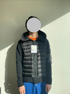 Svart pufferjacka från Moncler - Snygg svart pufferjacka från Moncler med quiltad front och huva. Jackan har stickade ärmar och ribbade muddar samt en ficka med dragkedja på ena ärmen. Klassisk Moncler-logga på armen och dragkedja framtill. Perfekt för kalla vinterdagar.