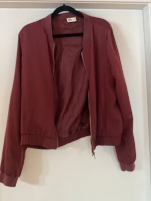 Vinröd bomberjacka från C&A - Snygg vinröd bomberjacka från C&A med klassisk rak passform och ribbade muddar vid ärmslut och midja. Jackan har dragkedja framtill och är tillverkad i ett lätt tyg som passar perfekt till lager på lager. Enkel och stilren design utan synliga fickor.