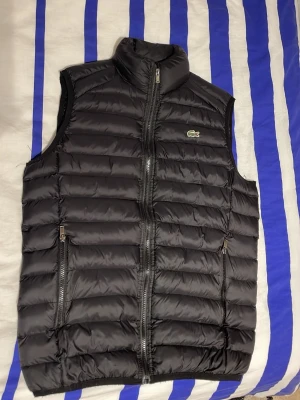 Svart dunväst från Lacoste - Snygg svart dunväst från Lacoste med klassisk krokodillogga på bröstet. Västen har hög krage, två dragkedjeförsedda fickor och quiltad design. Perfekt för lager-på-lager och ger en sportig vibe till din outfit.