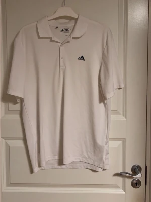 Vit pikétröja från Adidas - Klassisk vit pikétröja från Adidas med korta ärmar och krage. Tröjan har två knappar vid halsen och en liten Adidas-logga broderad på bröstet. Perfekt för golf eller andra sportaktiviteter.