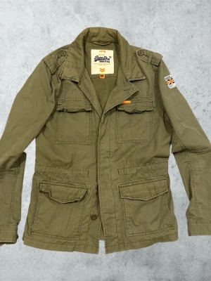 Olivgrön parkajacka från Superdry - Säljer en olivgrön parkajacka från Superdry med fyra stora fickor framtill, axelklaffar och patch med brittisk flagga på ärmen. Jackan har klassisk krage och stängs med knappar. Perfekt för dig som gillar utility-stil. Mått: Bredd; 40 cm. Längd; 68 cm. Ärm; 62 cm.