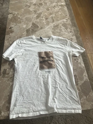 Vit Burberry t-shirt med beige patch - Säljer en vit t-shirt från Burberry med en stor beige patch på bröstet och svart Burberry-tryck. T-shirten har klassisk passform och korta ärmar. Materialet känns mjukt och är troligtvis bomull. Perfekt för dig som gillar exklusiva streetwear-plagg.