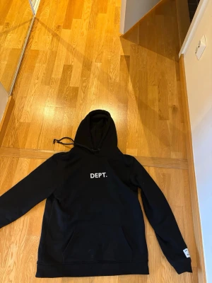 Svart hoodie från Gallery Dept. - Svart hoodie från Gallery Dept. med vit text 'DEPT.' framtill och 'GALLERY DEPT.' på ryggen. Klassisk modell med huva och dragsko samt känguruficka. Perfekt för en avslappnad streetwear-look.