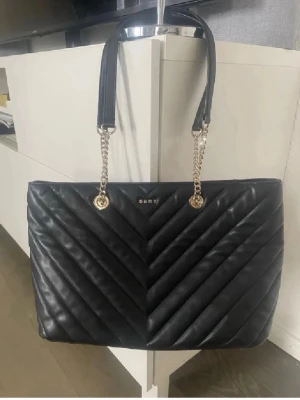 Svart quiltad handväska från DKNY - Snygg svart handväska från DKNY i quiltat läder med guldfärgade kedjedetaljer på handtagen. Väskan har en klassisk rektangulär form och diskret DKNY-logga framtill. Perfekt för dig som gillar stilrena och trendiga accessoarer.