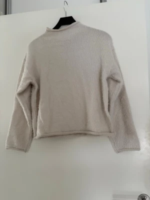 Beige fluffig stickad tröja - Mjuk och fluffig stickad tröja i beige färg med lång ärm och hög hals. Tröjan har en enkel och avslappnad passform, perfekt för att hålla sig varm under kyliga dagar. Materialet är lent och ger en cozy känsla. Oanvänd strl M