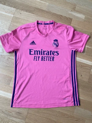 Rosa Real Madrid tröja Adidas M - Snygg rosa Real Madrid matchtröja från Adidas med mörkblå detaljer och logga. Tröjan har korta ärmar, rund hals och AEROREADY-material som andas. Tryck med 'Emirates Fly Better' på bröstet och klassiska Adidas-ränder på sidorna.