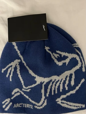 Blå mössa från Arc'teryx - Snygg blå mössa från Arc'teryx med vitt grafiskt fossil-mönster och broderad logga framtill. Mössan är stickad och har en klassisk, rund form som passar perfekt för kalla dagar. Perfekt för dig som gillar outdoor och streetstyle.