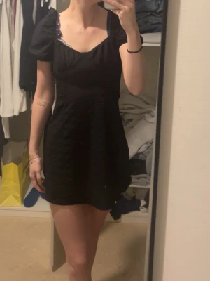 Svart kort klänning med puffärm - Snygg svart kort klänning med puffärmar och rundad urringning. Klänningen har en figurnära passform upptill och faller ut något nedtill. Perfekt för dig som gillar en enkel men trendig stil.