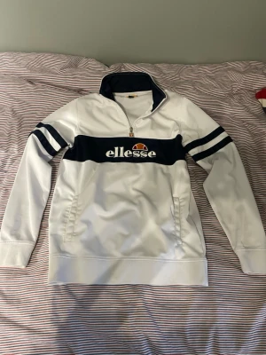 Vit och marinblå Ellesse sweatshirt - Säljer en vit sweatshirt från Ellesse med marinblått band och logga över bröstet. Tröjan har halv dragkedja, ståkrage och två fickor framtill. Marinblå ränder på ärmarna ger en sportig look. Perfekt för dig som gillar streetwear.