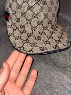 Beige Gucci keps med GG-mönster - Säljer en beige keps från Gucci med klassiskt GG-monogram i svart över hela kepsen. På sidan finns en ikonisk röd och marinblå rand. Kepsen har böjd skärm och är tillverkad i canvasmaterial. Perfekt för dig som vill ha en stilren och lyxig accessoar.