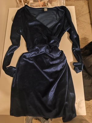 Mörkblå sammetsklänning omlott - Snygg mörkblå omlottklänning i mjuk sammet med lång ärm och v-ringning. Klänningen har en markerad midja och elegant glans i tyget, perfekt för dig som vill sticka ut. Passar till många olika tillfällen och ger en lyxig känsla.