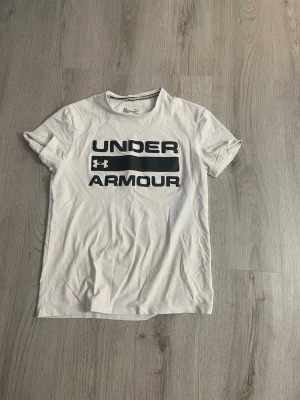 Vit Under Armour t-shirt - Snygg vit t-shirt från Under Armour med stor svart logga och text på bröstet. Klassisk rund halsringning och korta ärmar. Tillverkad i mjuk bomull som känns skön mot huden. Perfekt för en sportig och avslappnad stil. Har en liten fläkt