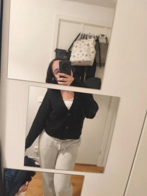 Svart stickad kofta med v-ringning - Säljer en svart stickad kofta med v-ringning och stora knappar framtill. Koftan har en avslappnad passform och långa ärmar, perfekt att slänga över en topp. Materialet ser mjukt och mysigt ut, och modellen är klassisk och enkel att matcha.