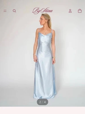 Ljusblå långklänning i satin By Stina - Elegant ljusblå långklänning från By Stina i glansigt satinmaterial. Klänningen har smala axelband, v-ringning och öppen rygg med snörning. Perfekt för dig som vill sticka ut med en stilren och modern look.