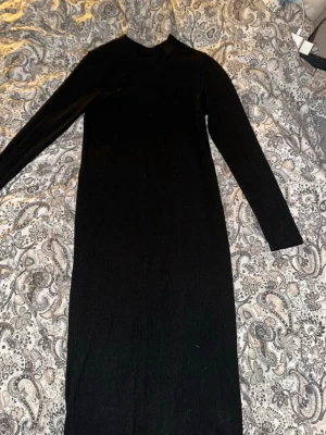 Svart långärmad ribbad klänning - En svart, långärmad klänning med ribbad struktur och slits nedtill på baksidan. Klänningen har en enkel, rak passform och hög hals. Perfekt för dig som gillar stilrena plagg. Den är stretchig 