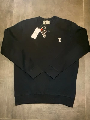 Svart sweatshirt från Ami Paris - Stilren svart sweatshirt från Ami Paris med rund halsringning och ikonisk liten vit Ami-de-Coeur-logga broderad på bröstet. Tillverkad i mjuk bomull som känns skön mot huden. Perfekt för en clean och avslappnad look.