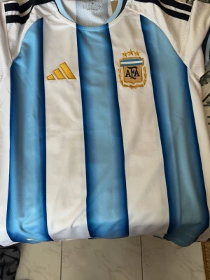 Argentina matchtröja Adidas blåvit - Säljer en officiell Argentina fotbollströja från Adidas med klassiska blå och vita vertikala ränder, guldfärgade Adidas-logga och AFA-emblem på bröstet. Tröjan har svarta detaljer på axlarna och siffran 1893 i nacken. Perfekt för fotbollsfans!
