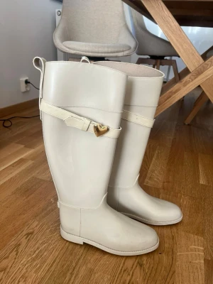 Vita höga stövlar med hjärtdetalj - Snygga vita höga love moschino stövlar i gummi med dekorativt spänne och guldfärgat hjärta på sidan. Stövlarna har rund tå, låg klack och dragflikar upptill. Perfekta för regniga dagar och ger en clean look till din outfit.