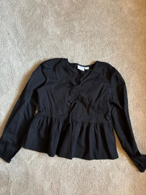 Svart blus med rosetter från Gina i storlek 146 / 152 - Svart blus med långa ärmar och v-ringning. Blusen har en markerad midja med knytband framtill och lätt rynkad nederdel för en avslappnad look. Tillverkad i ett mjukt, vävt tyg som känns skönt mot huden.