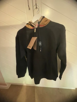 Svart hoodie med Burberry-mönstrad huva - Säljer en svart hoodie från Burberry med klassisk beige, röd och svart rutmönstrad huva. Hoodien har dragsko i huvan och ribbade muddar vid ärmar och nederkant. Perfekt för dig som vill ha en stilren men ändå lyxig look.