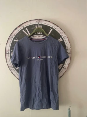 Blå t-shirt från Tommy Hilfiger - Snygg blå t-shirt från Tommy Hilfiger med klassisk logga och texten 'EST. 1985' på bröstet. T-shirten har rund halsringning och korta ärmar. Tillverkad i mjuk bomull som känns skön mot huden. Perfekt till jeans eller shorts.