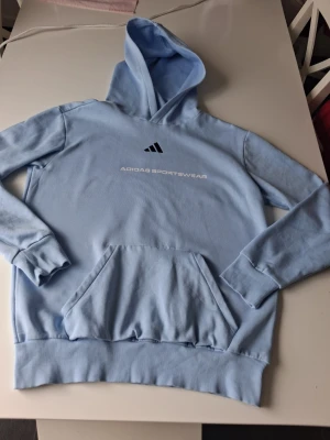 Ljusblå hoodie från Adidas - Snygg ljusblå hoodie från Adidas med trycket 'ADIDAS SPORTSWEAR' på bröstet och klassiska Adidas-loggan ovanför. Hoodien har en stor känguruficka framtill, ribbade muddar och huva. Perfekt för en avslappnad och sportig stil. Varan är i mycket bra skick i storlek M. Hör av dig vid frågor