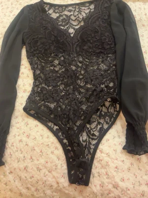 Svart spetsbody med långa ärmar - Snygg svart body i spets med långa, transparenta ärmar och djup v-ringning. Kroppen är helt i blommig spets och ärmarna är i tunnare tyg med resår vid handlederna. Perfekt för att lyfta vilken outfit som helst.