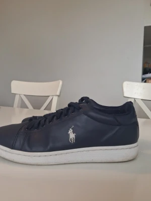 Mörkblå Polo Ralph Lauren sneakers - Snygga mörkblå sneakers från Polo Ralph Lauren med klassisk vit logga på sidan och POLO-text bak på hälen. Skorna har lågt skaft, vita sulor och är gjorda i slätt lädermaterial. Perfekta för en clean och stilren look. Varan är i mycket bra skick i storlek 43