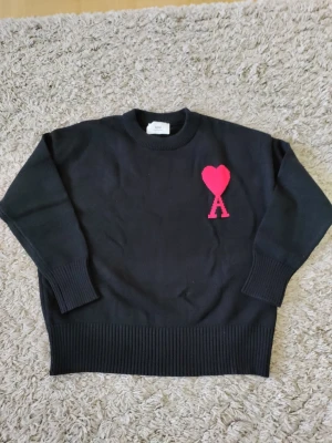 Svart stickad tröja från Ami Paris - Säljer en svart stickad tröja från Ami Paris med den ikoniska röda hjärtloggan och bokstaven A på bröstet. Tröjan har rund hals, ribbade muddar och nederkant. Perfekt för dig som gillar stilrena plagg med en twist.